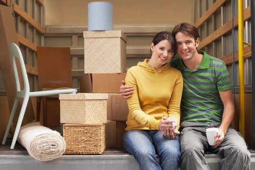 A young couple is sitting inside the open back of a residential removals van, surrounded by various packed moving boxes made of cardboard and woven materials, with some boxes stacked behind them. The woman, wearing a yellow hoodie and blue jeans, is smiling and holding a cup, while the man, dressed in a green and purple striped polo shirt and light grey trousers, is also smiling and holding a white mug. In front of them, a rolled-up rug and a light-colored chair with a fabric seat are visible, with a small cylindrical container placed on top of a beige storage box. The interior of the van features wooden panels and shelving, indicating a house or apartment move, and soft natural lighting suggests daytime. This scene reflects a successful move completed by Uxbridge Man with Van, a house removals service operating in the Uxbridge area, capturing a moment of satisfaction and teamwork during a household relocation.